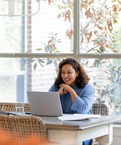 The mid adult woman sits in the dining room and uses her laptop for a video call with her family.